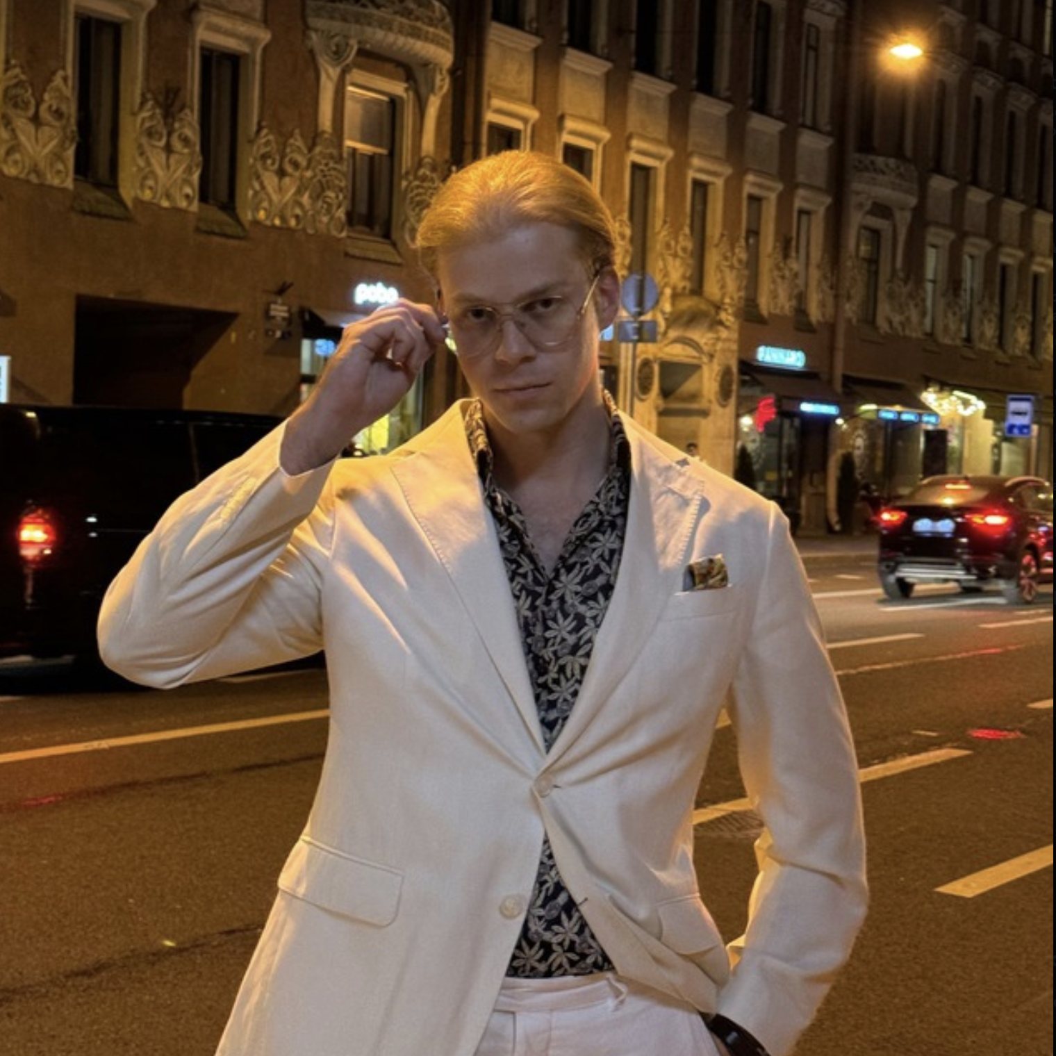 A man in a white suit adjusting his glasses on a city street at night
